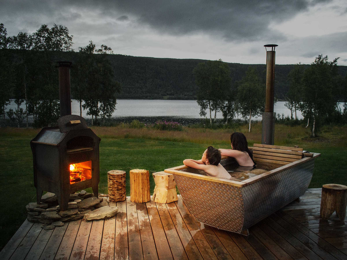 "Två personer badar i ett uppvärmt utomhusbadkar och tittar in i en flammande eld bredvid."